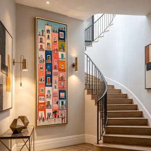 Op de afbeelding: Een kleurrijk, ingelijst kunstwerk dat een straatbeeld weergeeft met gebouwen in verschillende kleuren, waaronder roze, blauw en oranje. Het kunstwerk hangt aan een lichtgrijze muur, naast een trap met houten treden en een zwarte balustrade.