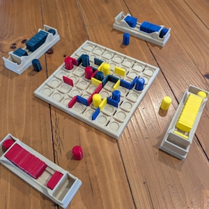 May include: A board game with a beige grid and colourful pieces. The game includes red, yellow, and blue rectangular and cylindrical pieces, along with four beige trays to hold the pieces. The game is set on a wooden surface.
