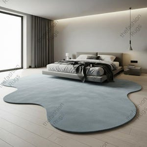 Può includere: Un grande tappeto azzurro con una forma organica irregolare simile a una nuvola, caratterizzato da bordi morbidi e arrotondati. Il tappeto è posizionato su un pavimento in legno chiaro in una camera da letto, sotto un letto a piattaforma.