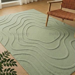 Op de afbeelding: Een saliegroen vloerkleed met een verhoogd, vloeiend, golvend patroon. Het rechthoekige vloerkleed heeft de tekst "thefashion Rugs" in de hoeken. Op de achtergrond staat een houten stoel met een geweven zitting.