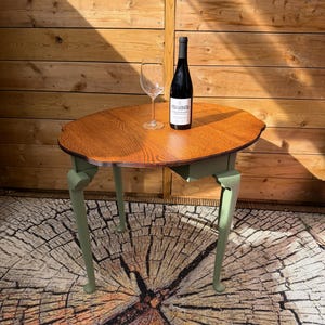 May include: A small, round wooden table with a brown tabletop and green legs. A bottle of wine and a wine glass sit on the table. The table is placed on a rug with a tree trunk design.