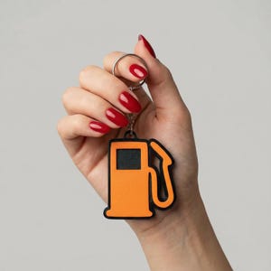 Könnte beinhalten: Orangener und schwarzer Tankstellen-Schlüsselanhänger. Der Schlüsselanhänger hat ein detailliertes Design einer Zapfsäule mit schwarzer Umrandung. Der Schlüsselanhänger wird von einer Hand mit rotem Nagellack gehalten.
