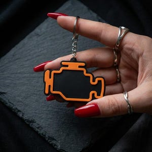 Peut inclure: Porte-clés en forme de moteur noir et orange. Le porte-clés a un centre noir avec un contour orange et est attaché à une chaîne et un anneau argentés. Le fond est sombre.