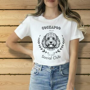 Könnte beinhalten: Weißes T-Shirt mit einem schwarz-weißen Cockapoo-Hundedesign. Der Text lautet "Cockapoo Hazel Social Club EST 2008", mit einem Lorbeerkranz um den Kopf des Hundes. Ein lässiges Kleidungsstück.