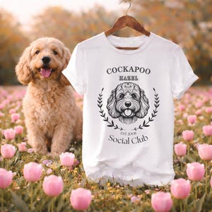 Può includere: Maglietta bianca con una grafica nera di un cane Cockapoo e la scritta "Cockapoo Hazel Social Club Est 2008". Un cane Cockapoo dorato è seduto accanto alla maglietta in un campo di fiori rosa.