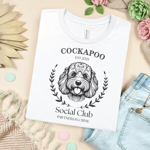 Könnte beinhalten: Weißes T-Shirt mit schwarzer Grafik eines Cockapoo-Hundes, Lorbeerkranz und Text "Cockapoo Est 2025" und "Social Club Pawtners in Crime". Das Shirt hat einen weißen Kragen.