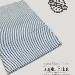 Może przedstawiać: Złożony kawałek białej tkaniny z niebieskimi poziomymi paskami. Tkanina jest oznaczona jako "100% Bawełna" i "Hand Block Print Rapid Print Pure Cotton Fabric".