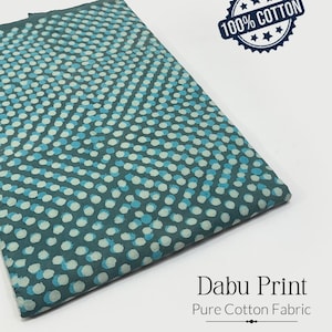 May include: Folded teal fabric with a dotted pattern in light blue and white. The fabric is labelled "Dabu Print" and "Pure Cotton Fabric." A blue and white circular stamp reads "100% Cotton."
