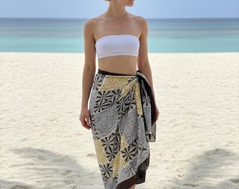 Puede incluir: Una mujer con un top blanco y un pareo estampado en una playa de arena. El pareo presenta un diseño geométrico en negro, blanco y amarillo. El océano y el cielo son visibles en el fondo.
