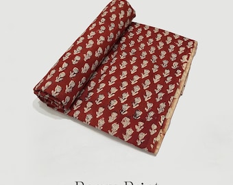 May include: A folded and rolled piece of Bagru Print pure cotton fabric. The fabric is a deep red color with a repeating pattern of off-white floral designs. The words "100% Cotton" are printed on a circular stamp.
