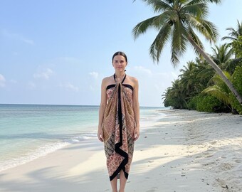 May include: A patterned sarong in shades of brown, black, and cream, worn on a beach. The sarong features a geometric design and is draped over the shoulders. The background includes a sandy beach, turquoise water, and palm trees under a blue sky.