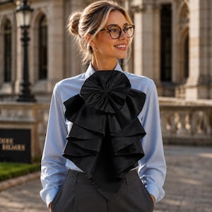 May include: A light blue long-sleeved button-down shirt featuring a large, black, multi-layered fabric ruffle attached to the front. The ruffle is circular at the top and cascades down in wide, pleated folds, creating a prominent decorative accent.