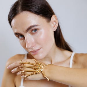 May include: A gold-colored skeleton hand bracelet with articulated fingers and a wrist cuff. The jewelry piece is worn on a wrist and extends to the fingers, creating a unique and gothic accessory. The bracelet is made of metal.