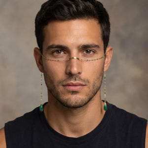 Puede incluir: Una cadena facial dorada con un diseño delicado. La cadena presenta pequeñas cuentas y dos colgantes en forma de lágrima de color verde esmeralda. La cadena facial se usa sobre el puente de la nariz y cae a ambos lados de la cara.