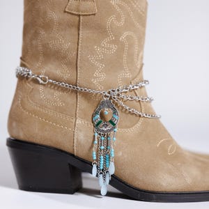 Op de afbeelding: Een zilverkleurige laarketting met een decoratieve hanger met turquoise en groene accenten en bungelende blauwe kralen. De ketting wordt getoond op een bruin suède cowboylaars met decoratieve stiksels.