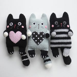 May include: Three handmade crochet cat dolls in black, grey, and black and white stripes. Each cat has a heart-shaped design on its chest, with pink polka dots on the black cat. The cats have button eyes and pink cheeks.