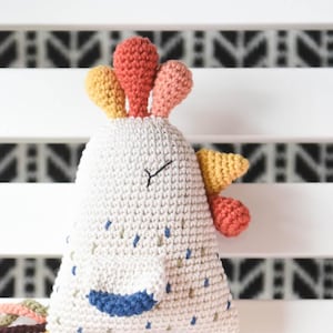 Puede incluir: Un juguete de gallo de crochet con cuerpo blanco, patas marrones y cola de colores. El gallo tiene un pico amarillo, una cresta naranja y amarilla y ojos azules.