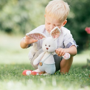 May include: A young child sits on the grass holding a crocheted bunny toy. The bunny is white with blue overalls and a brown button. The bunny's ears are pink and white with a floral pattern.
