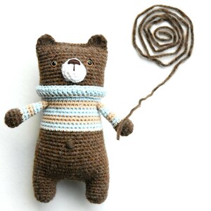 Puede incluir: Un oso de crochet marrón que lleva un suéter a rayas azul y beige sostiene una bola de hilo marrón en su pata.
