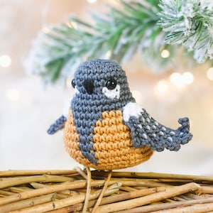 Op de afbeelding: Een gehaakt vogelsieraad van grijs, wit en oranje garen. De vogel zit op een gevlochten takkenkrans.