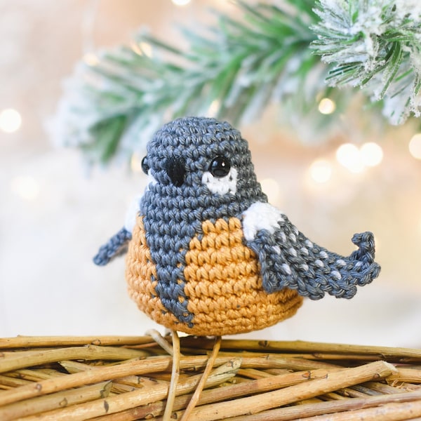 Puede incluir: Un adorno de pájaro de crochet hecho con hilo gris, blanco y naranja. El pájaro está posado en una corona de ramas trenzadas.