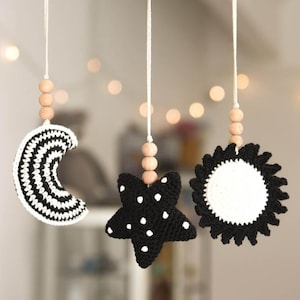 May include: A handmade baby mobile featuring a wooden hoop with three hanging crochet shapes: a crescent moon, a star, and a sun. The moon has black and white stripes, the star is black with white dots, and the sun is white with a black border.