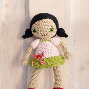 Puede incluir: Una muñeca de crochet con cabello negro, ojos verdes y un vestido rosa y verde. La muñeca lleva zapatos rojos y está sentada sobre una superficie de madera.