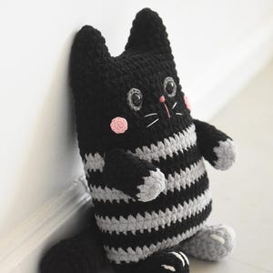 Pode incluir: Boneca de gato de croch&ecirc; listrada em preto e cinza feita &agrave; m&atilde;o. O gato tem corpo, cabe&ccedil;a e cauda pretos, com torso e bra&ccedil;os listrados em cinza e preto. Possui bochechas rosadas, olhos cinzentos e bigodes brancos. O gato est&aacute; encostado em uma parede branca.