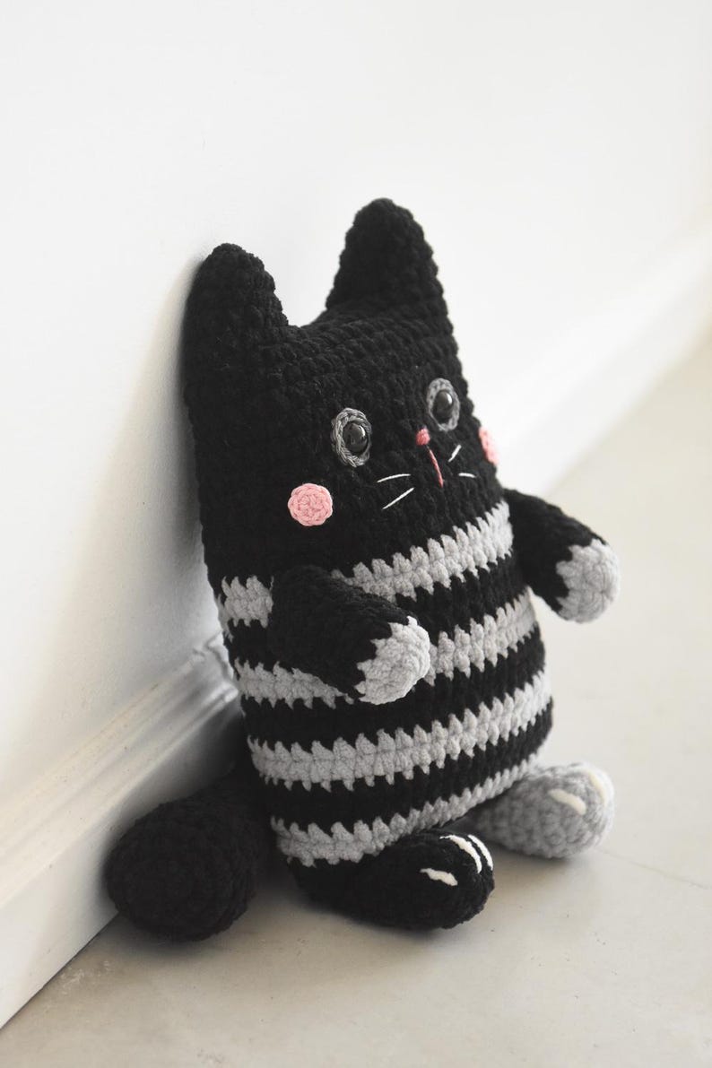 Pode incluir: Boneca de gato de croch&ecirc; listrada em preto e cinza feita &agrave; m&atilde;o. O gato tem corpo, cabe&ccedil;a e cauda pretos, com torso e bra&ccedil;os listrados em cinza e preto. Possui bochechas rosadas, olhos cinzentos e bigodes brancos. O gato est&aacute; encostado em uma parede branca.
