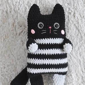 Pode incluir: Um gato de pel&uacute;cia de croch&ecirc; feito &agrave; m&atilde;o, com listras brancas e pretas, bochechas rosa e nariz rosa. O gato tem olhos grandes e redondos e bigodes brancos. A cauda e as patas s&atilde;o pretas, e os p&eacute;s cinzentos. A pel&uacute;cia est&aacute; sobre uma superf&iacute;cie cinzenta texturizada.