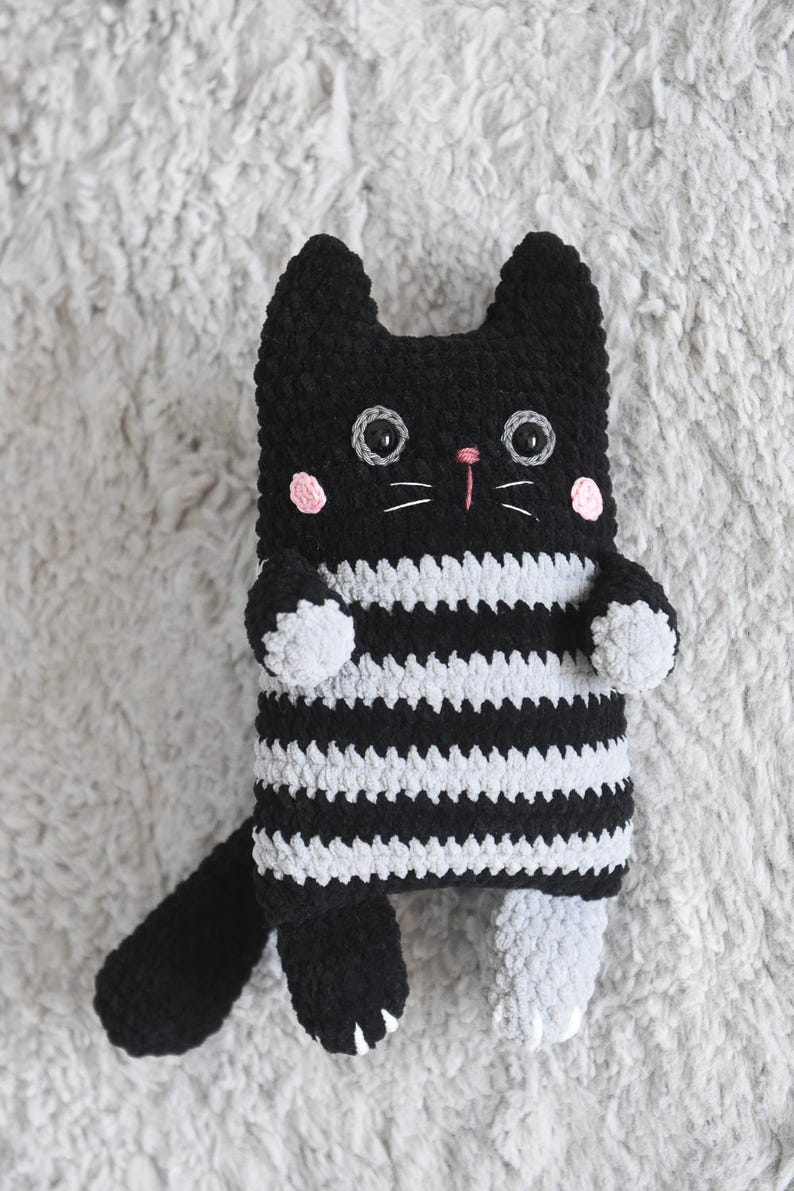 Pode incluir: Um gato de pel&uacute;cia de croch&ecirc; feito &agrave; m&atilde;o, com listras brancas e pretas, bochechas rosa e nariz rosa. O gato tem olhos grandes e redondos e bigodes brancos. A cauda e as patas s&atilde;o pretas, e os p&eacute;s cinzentos. A pel&uacute;cia est&aacute; sobre uma superf&iacute;cie cinzenta texturizada.