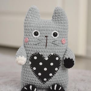Pode incluir: Boneca de gato de croch&ecirc; cinza feita &agrave; m&atilde;o com um patch preto em forma de cora&ccedil;&atilde;o com bolinhas brancas. O gato tem olhos pretos e brancos, bochechas rosadas e patas pretas. A boneca est&aacute; em uma superf&iacute;cie cinza.