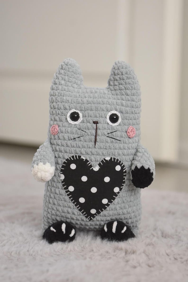 Pode incluir: Boneca de gato de croch&ecirc; cinza feita &agrave; m&atilde;o com um patch preto em forma de cora&ccedil;&atilde;o com bolinhas brancas. O gato tem olhos pretos e brancos, bochechas rosadas e patas pretas. A boneca est&aacute; em uma superf&iacute;cie cinza.