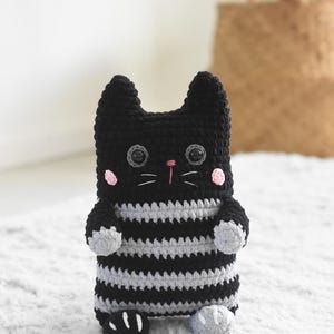 Pode incluir: Boneca de gato de croch&ecirc; preta e cinza feita &agrave; m&atilde;o. O gato tem olhos grandes, bochechas rosadas e nariz rosa. O corpo &eacute; listrado com fio cinza e preto. O gato tem pequenas patas e orelhas cinzentas.