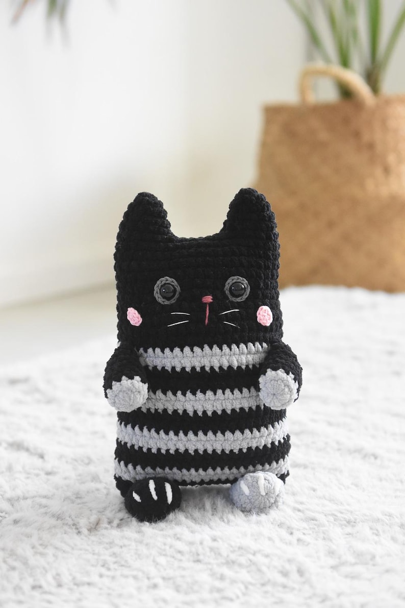 Pode incluir: Boneca de gato de croch&ecirc; preta e cinza feita &agrave; m&atilde;o. O gato tem olhos grandes, bochechas rosadas e nariz rosa. O corpo &eacute; listrado com fio cinza e preto. O gato tem pequenas patas e orelhas cinzentas.