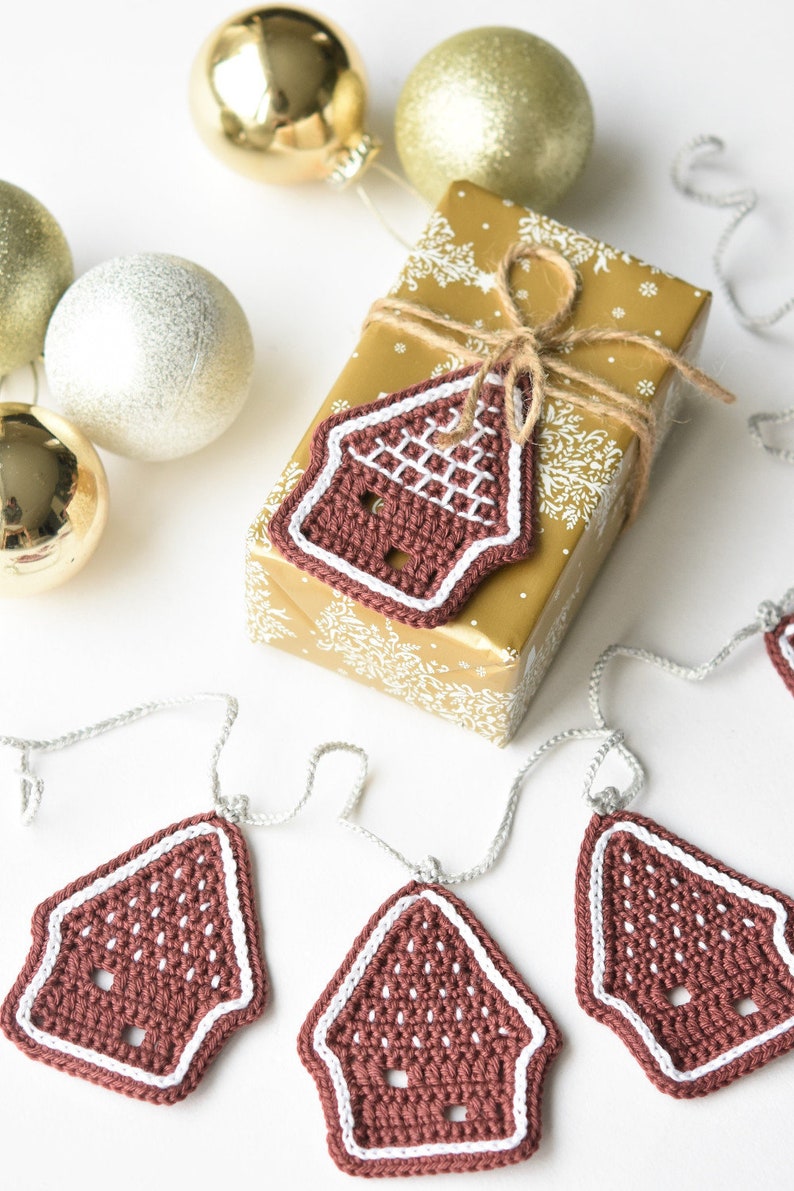 May include: A brown crocheted gingerbread house ornament with white trim hangs from a silver string. The ornament is part of a garland of gingerbread houses. The garland is displayed on a white surface with a gold gift box wrapped with twine and a gingerbread house ornament.