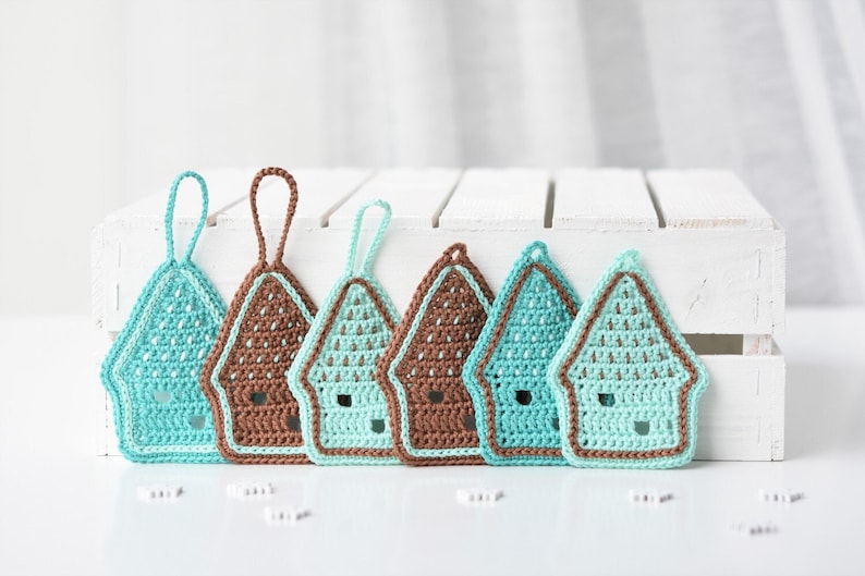 May include: Six crocheted house ornaments in brown and teal colours. The ornaments are hanging from a white wooden crate.