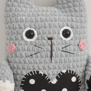 Pode incluir: Um gato de pel&uacute;cia de croch&ecirc; cinza feito &agrave; m&atilde;o com uma aplica&ccedil;&atilde;o em forma de cora&ccedil;&atilde;o preto com bolinhas brancas. O gato tem olhos pretos, bochechas rosadas e bigodes bordados. As orelhas est&atilde;o em p&eacute;.