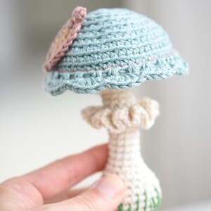 May include: A crocheted mushroom toy with a light blue cap and a white stem. The cap has a pink flower detail and the stem has green accents.
