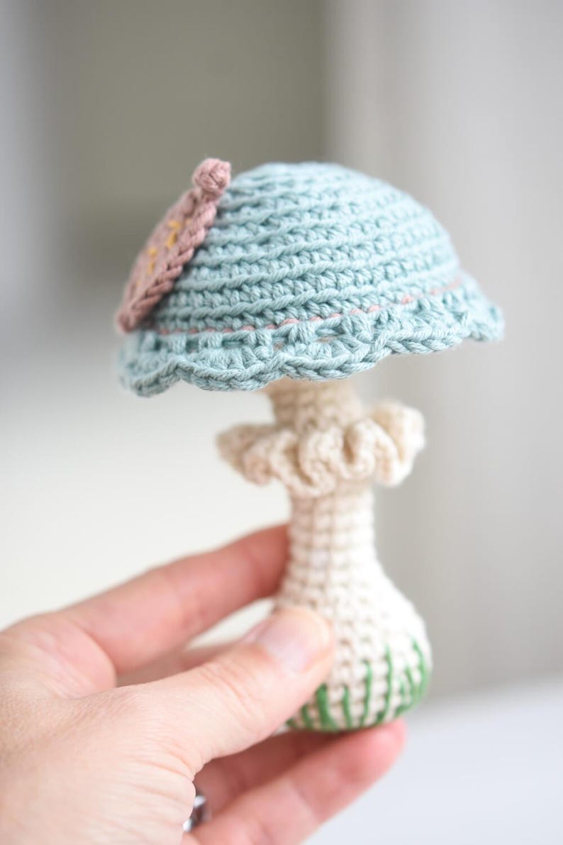 May include: A crocheted mushroom toy with a light blue cap and a white stem. The cap has a pink flower detail and the stem has green accents.