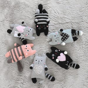 Pode incluir: Uma cole&ccedil;&atilde;o de brinquedos de gato feitos &agrave; m&atilde;o em croch&ecirc; em v&aacute;rias cores e padr&otilde;es. Os gatos s&atilde;o cinza, preto, rosa e azul, com detalhes listrados e em forma de cora&ccedil;&atilde;o. Cada gato tem um design &uacute;nico.