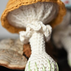 May include: A crocheted mushroom toy with a yellow hat and a green and white stem. The mushroom is sitting on a brown surface.