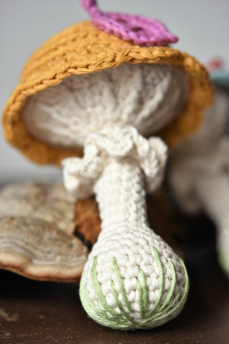 May include: A crocheted mushroom toy with a yellow hat and a green and white stem. The mushroom is sitting on a brown surface.