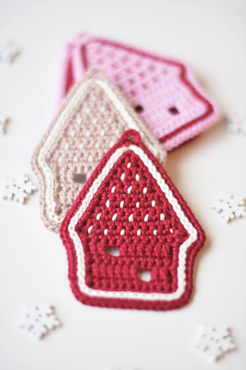 May include: Three crocheted gingerbread house ornaments in red, pink and beige. The ornaments are decorated with white crocheted trim.