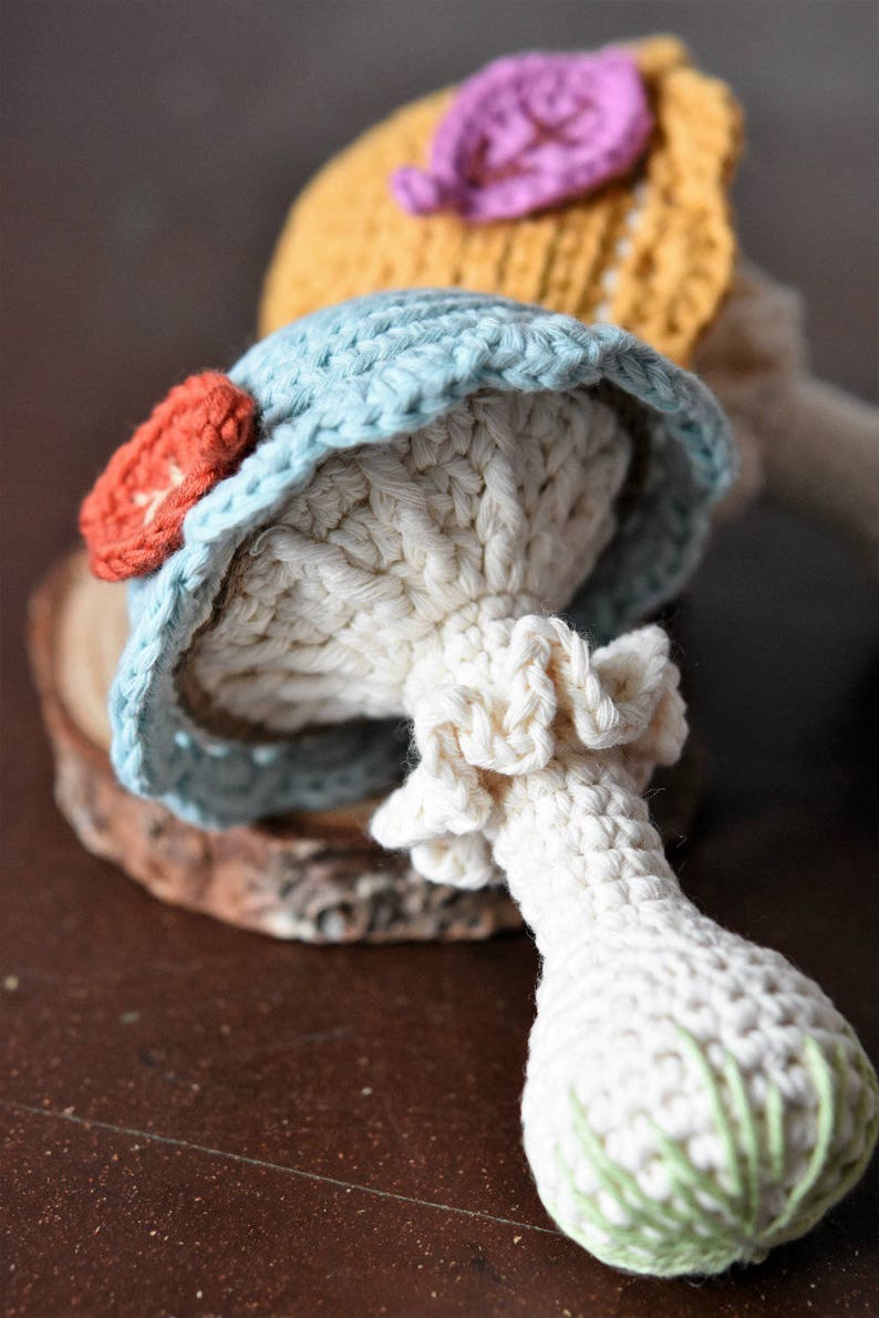 May include: Two crocheted mushroom toys. One is blue with a red flower and the other is yellow with a purple flower. Both have white stems.