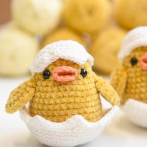 Puede incluir: Dos pollitos amarillos de crochet en cáscaras de huevo blancas. Los pollitos tienen ojos negros y picos naranjas.