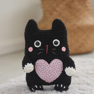 Pode incluir: Boneca de gato de croch&ecirc; preta feita &agrave; m&atilde;o com olhos brancos, bochechas rosadas e um patch em forma de cora&ccedil;&atilde;o rosa no peito. O gato tem patas brancas e est&aacute; em p&eacute; sobre um tapete de cor clara. O gato tem orelhas pequenas e um nariz rosa.