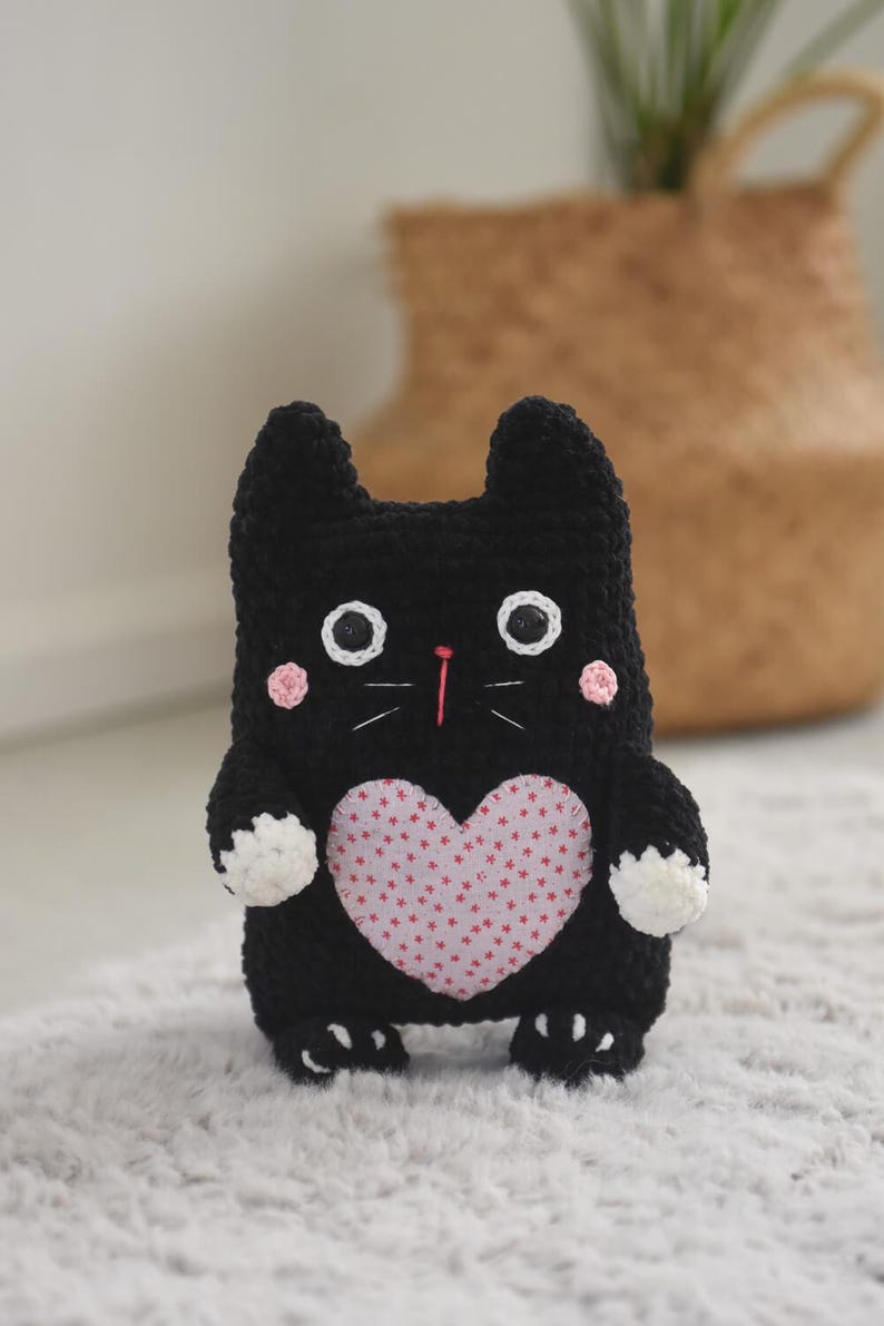 Pode incluir: Boneca de gato de croch&ecirc; preta feita &agrave; m&atilde;o com olhos brancos, bochechas rosadas e um patch em forma de cora&ccedil;&atilde;o rosa no peito. O gato tem patas brancas e est&aacute; em p&eacute; sobre um tapete de cor clara. O gato tem orelhas pequenas e um nariz rosa.