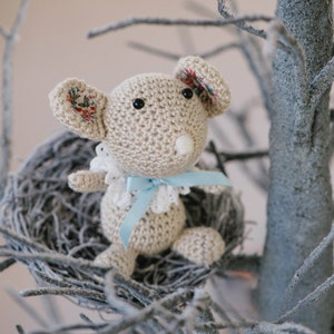 May include: A crocheted mouse toy with a blue ribbon and floral fabric ears sits in a gray nest on a branch of a faux snow-covered tree.