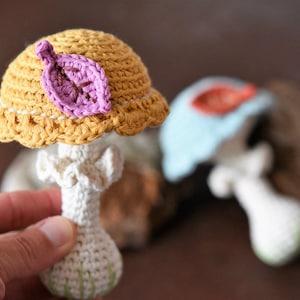 May include: A crocheted mushroom toy with a yellow cap and a pink leaf. The mushroom has a white stem and is made with a soft yarn.